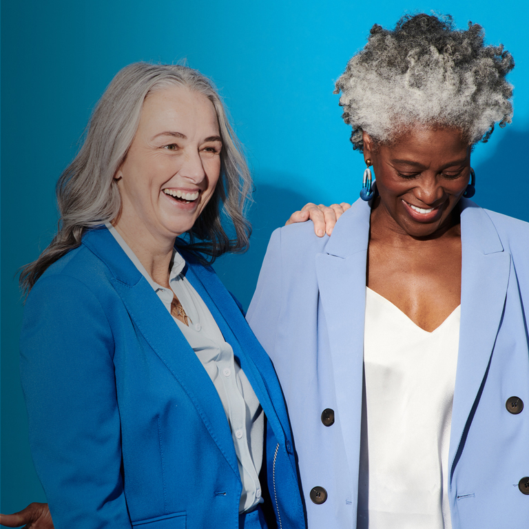 Two mature professional women, one black and one white, dressed in shades of blue, smile and display friendship and professionalism