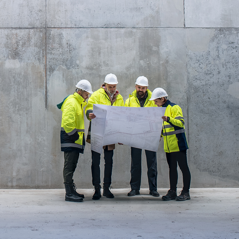 A group of engineers standing against concrete wall on construction site, holding blueprints. Copy space.