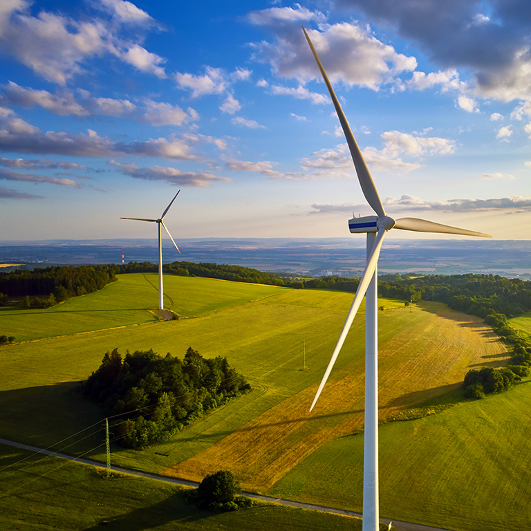 Aerial view of wind turbine farm. Wind power plants in green landscape against sunset sky with clouds. Aerial, drone inspection of wind turbine.