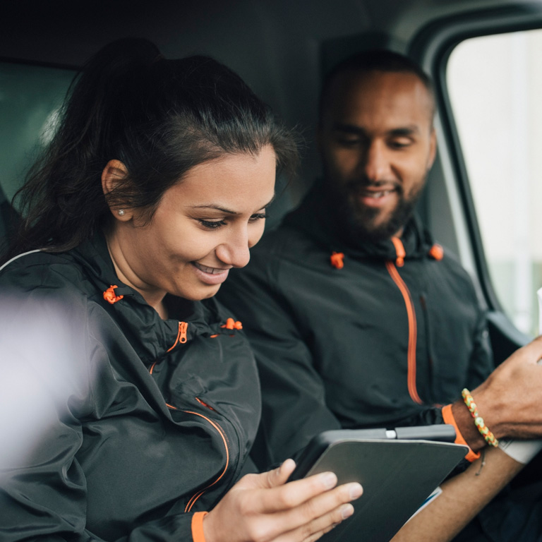 Two delivery workers in a van smiling and looking at a tablet while one hols a parcel
