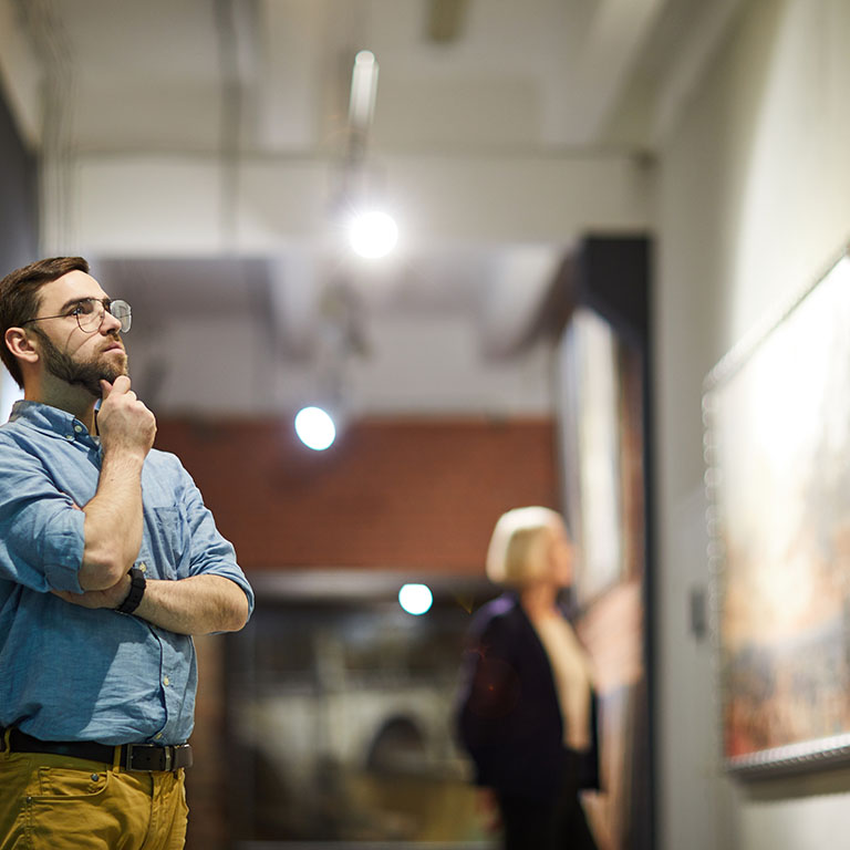 Portrait of pensive bearded man looking at paintings standing in art gallery or museum, copy space