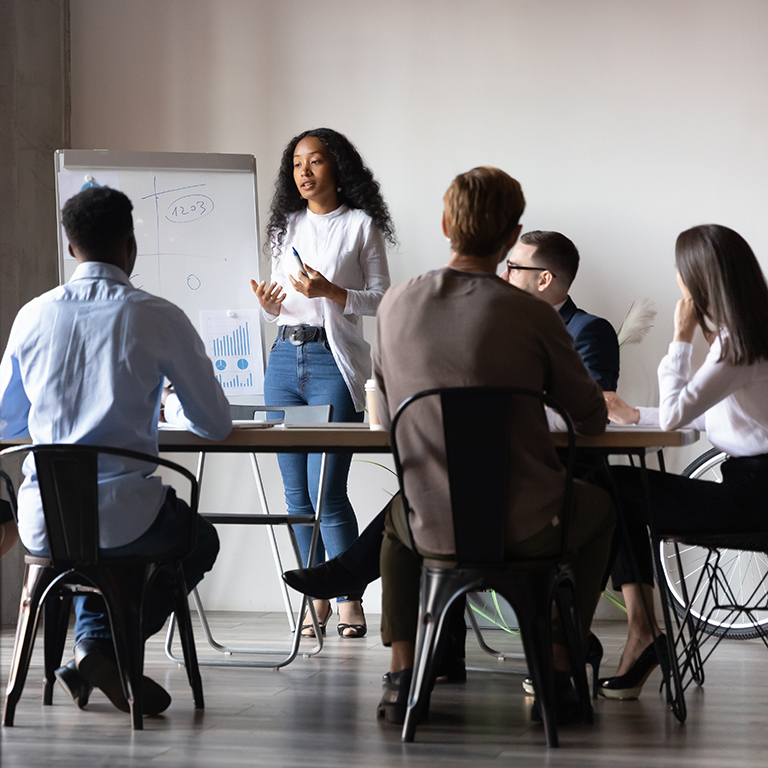 Six diverse businesspeople gathered in modern board room, different age ethnicity workers listen african woman coach tell about strategy, reporting using flip chart. Training and negotiations concept